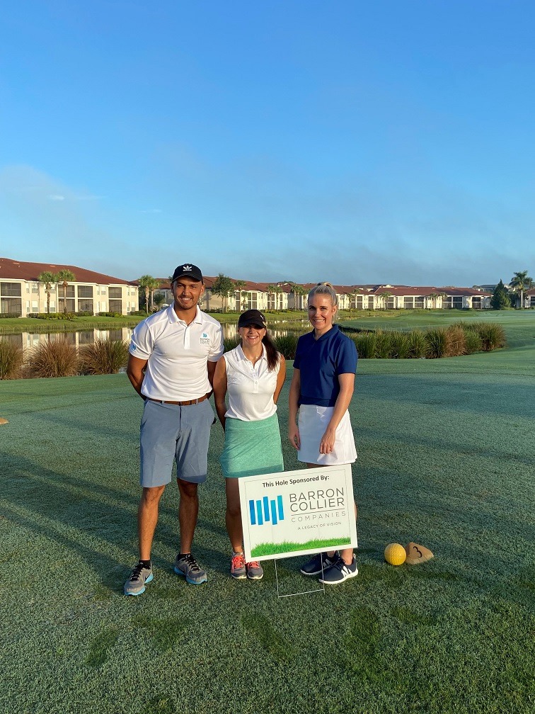 Three people standing on gold course close to Barron Collier Companies sign